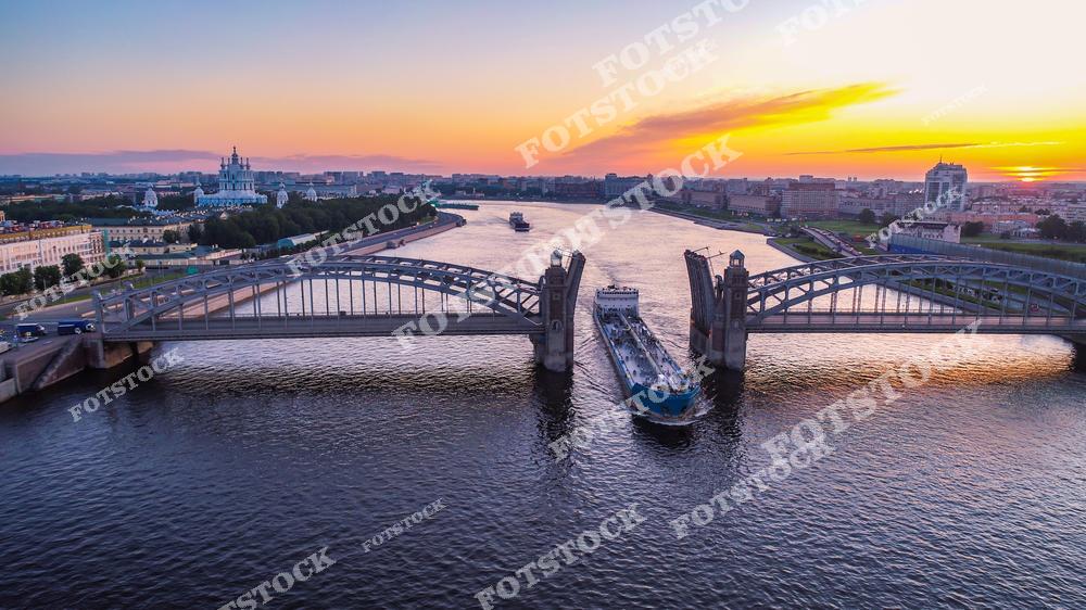 Панорамная фотография Санкт-Петербурга с высоты птичьего полета на закате. На снимке виден Большеохтинский мост, по которому движется транспорт, река Нева, а вдали — исторические здания и зеленые зоны города. Атмосфера вечернего города передается мягким светом заката и отражением в воде. Съемка с дрона позволяет оценить масштабы городской архитектуры, протяженность мостов и красоту городской панорамы с уникального ракурса. Фотография идеально подходит для публикаций о Петербурге, туристических материалов, фотостоков, оформления интерьеров и рекламных проектов, связанных с городскими пейзажами и архитектурой.